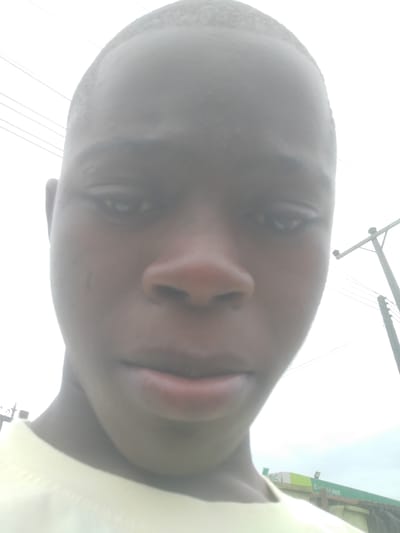 A close-up selfie of a young black man with short hair. The man is wearing a light-colored shirt. The photo is taken outdoors, with utility poles and power lines visible in the background. A building with a green and white sign is also partially visible. Based on the location of the building, this is in Owode-Elede, Nigeria.