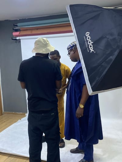 A photographer in a black outfit and a white bucket hat is taking a picture of two men. One man is wearing a blue robe, a gold watch, and a traditional Yoruba cap. The other man is wearing a yellow outfit. A large Godox softbox light is visible on the upper left. The scene is set up in a studio with white and grey backgrounds and multi-colored paper backdrops.