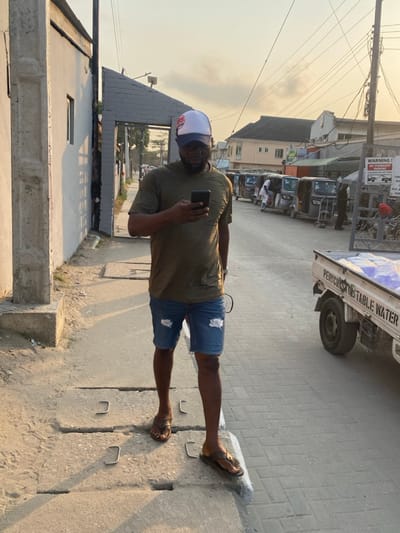 A man is standing on a concrete sidewalk, holding a phone and seemingly taking a selfie. He is wearing a light green t-shirt, ripped denim shorts, a baseball cap, and sandals. In the background, a street scene unfolds, with a vehicle carrying water, parked auto rickshaws, and buildings under a hazy sky. The setting appears to be an urban area.