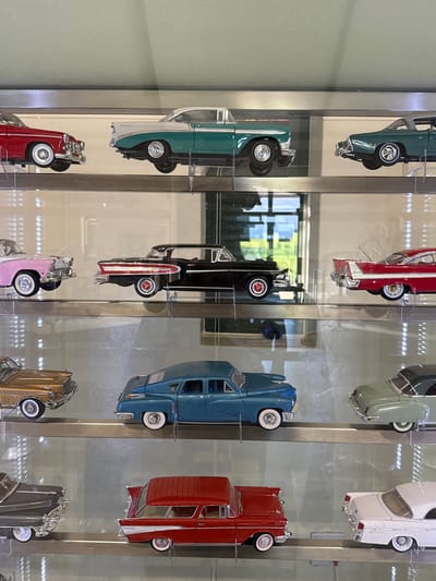 The image shows a display case containing several model cars. The cars are arranged vertically in rows, with a variety of colors and styles visible. The display case appears to be made of glass and metal, with the cars resting on clear shelves.

The reflections in the glass suggest the location is indoors, likely within a building or showroom. The location within Stellenbosch Local Municipality, South Africa is not identifiable from the visual data alone.