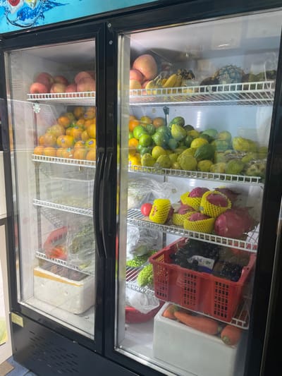 A commercial glass-door refrigerator, black-framed with two clear doors, is situated indoors, likely within a retail establishment in Dali City, China. The unit is filled with various fresh produce on multiple wire shelves.

The left door displays, from top to bottom:
*   Red apples on the uppermost shelf.
*   A large quantity of oranges or tangerines on the second shelf, some bearing small white stickers.
*   The third shelf contains produce in clear plastic bags, including green leafy items.
*   The bottom shelf holds a white polystyrene foam box containing a dark, solid item, possibly meat.

The right door displays, from top to bottom:
*   The uppermost shelf contains red apples, yellow bananas, and a whole pineapple.
*   The second shelf is densely packed with green and yellow-green fruits, including limes, possibly guavas, and other citrus varieties.
*   The third shelf features multiple red dragon fruits individually wrapped in yellow foam netting, a single red tomato, and green bitter gourds enclosed in clear plastic bags.
*   The bottom shelf holds a red plastic crate filled with dark grapes. Beneath the crate, a white polystyrene foam box contains orange carrots.

The background above the refrigerator features a light blue surface with a white and blue graphic depicting water, suggesting a beverage or water cooler is nearby. The floor beneath the refrigerator is light-colored. No people are visible in the frame.