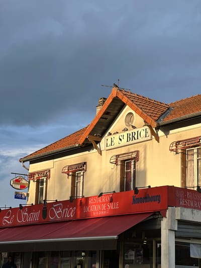 The image depicts the front of a building in Saint-Brice-sous-Forêt, France, identified as "Le Saint Brice," a restaurant and bar-brasserie. The building has a red-tiled roof and a light-colored façade with windows adorned with red metal balcony railings. A red awning stretches over the entrance, displaying the names "Le Saint Brice" and "Kronenbourg." There is a sign indicating "RESTAURANT LOCATION DE SALLE" with a telephone number. Additional signs include a "Kronenbourg" beer advertisement and an "FDJ" (Française des Jeux) sign. The sky above is overcast with dark clouds. There is evidence of additional storefronts to the right of this building.