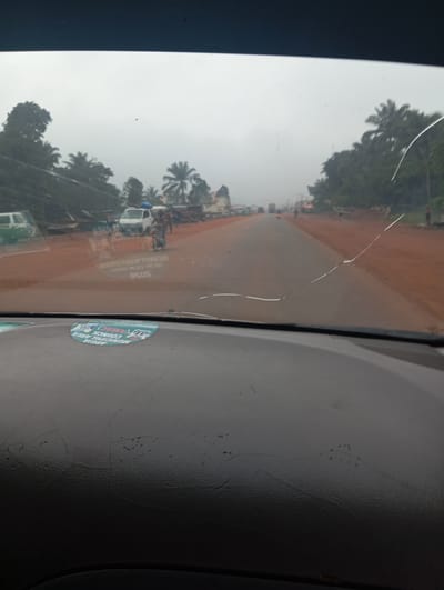A cracked windshield obscures the view of a street in Imilike, Nigeria. On the road, several vehicles are visible, including a white van, a motorcycle with a passenger, and other cars and trucks further down the road. The street is lined with trees and buildings on either side. The sky is overcast, and the overall lighting suggests a gloomy or rainy day. Two stickers are present on the windshield, one of which is partially obscured. The dashboard is visible in the foreground.