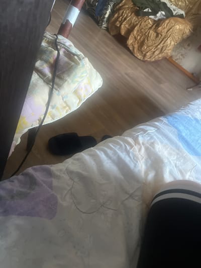 The image depicts an indoor scene, likely a bedroom, with a focus on a bed and the surrounding floor. A dark brown, possibly wooden furniture piece is visible on the left. The bed is covered with a quilt featuring a pattern of purple and white floral elements. A black slipper is placed on the wooden floor next to the bed. In the background, there's a crumpled golden-colored fabric and some clothing items. A portion of a person's leg, wearing black stockings with white stripes, is visible on the right side of the image, resting on the bed. The floor is made of light-colored wooden planks. The scene appears to be a domestic setting in Chemashikha, Russia.