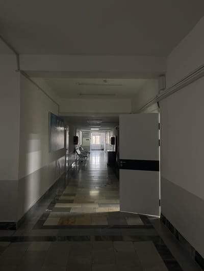 The image depicts an unoccupied, dimly lit hallway in a building located in Navoiy City, Uzbekistan. The perspective is a straight-on view down a long corridor. The ceiling is white, featuring multiple rectangular fluorescent light fixtures, some illuminated, and a smoke detector. White conduit runs along the upper perimeter of the walls. The walls are painted white, with a darker grey dado rail covering the lower portion.

On the left wall, a large, rectangular poster or information board is mounted. Further down the left, a set of three connected metal waiting chairs with black seats are positioned against the wall. Beyond these chairs, a dark, low cabinet or desk is partially visible.

On the right side of the hallway, a white door with a dark horizontal stripe on its lower half stands ajar, revealing a thin vertical line of bright light emanating from the room beyond. Further down the right wall, a small, dark red box, likely a fire alarm or extinguisher, is mounted.

The floor is tiled with a multi-patterned design consisting of rectangular tiles in shades of grey, beige, and dark brown. Sunlight streams from a large window at the far end of the hallway, creating reflections and bright patches on the polished floor. The window appears to have vertical blinds or bars. No people are present in the scene.