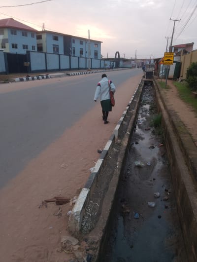 The image depicts a street scene in Alagbado, Nigeria. A person walks away from the camera on a sandy path next to a concrete drainage ditch filled with murky water and debris. The drainage ditch is bordered by a concrete curb with black and white stripes. To the left of the path is a paved road. In the background, several light blue buildings with a red-tiled roof line the street, enclosed by a light blue wall. A yellow and black speed hump warning sign is visible on a pole on the right side of the road. The sky is overcast with a soft, diffused light, suggesting it may be early morning or late afternoon.