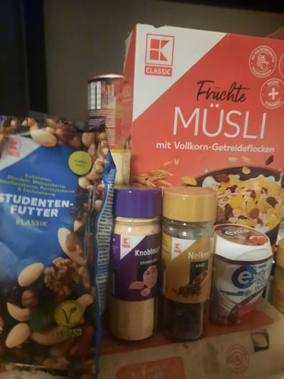 The image shows a collection of food items on a shelf. In the foreground, on the left, is a large blue bag labeled "STUDENTENFUTTER KLASSIK," which appears to be a mix of nuts and dried fruits, with a circular "VEGAN" sticker. To the right of the bag are two spice jars. The one on the left has a purple lid and is labeled "Knoblauch GRANULIERT." The one to its right has a yellow lid and is labeled "Nelken GANZ." Further to the right is a white container with blue and red labeling, partially obscured, which appears to be a dental product, possibly toothpaste, labeled "Extra Profi-Weiß." Behind these items, on the right side of the image, is a large red and white box of "Früchte MÜSLI mit Vollkorn-Getreideflocken." A portion of another item, possibly a cereal box or food packaging, is visible behind the muesli box, with a prominent "K CLASSIC" logo. The background is a dark, neutral color, suggesting a shelf or cupboard. The scene is consistent with items found in a grocery store or pantry, with no discernible landmarks indicating a specific location within Rosenheim, Germany. No people or actions are depicted in the image.