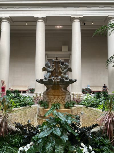 This photograph captures the interior courtyard of the National Gallery of Art in Washington, D.C. A classical-style fountain, adorned with two cherub sculptures, is the central focus of the scene. The fountain is surrounded by lush greenery and a shallow pool with a pebble-lined bottom. Tall, white columns support the building's architecture, creating a sense of grandeur and symmetry. Several visitors are present, with a woman in a red top walking past the fountain, and two individuals seated on a bench in the background. The lighting suggests it is daytime, possibly overcast or in a shaded area.