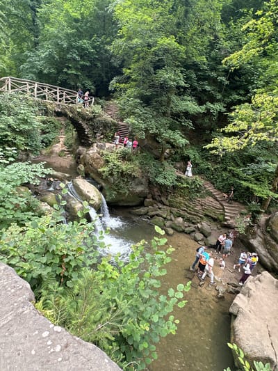 The image captures a view of the watermill of Waldbillig, Luxembourg, a picturesque natural setting with a small waterfall cascading into a shallow river. A stone bridge, partially covered in greenery, spans the river, with a wooden railing along its top. Stone steps lead up from the riverbank to the bridge, and further stairs continue up the hillside. Several people are present, scattered across the scene. On the bridge, a couple is walking. Further up the stairs, a group of people are gathered, some observing the surroundings. Near the river, closer to the waterfall, another group of people are wading in the water or standing on the rocks. A dog is visible on the stairs. The environment is lush and verdant, dominated by trees and vegetation, suggesting a tranquil, natural park or forest area. The lighting indicates it is daytime, with sunlight filtering through the trees.