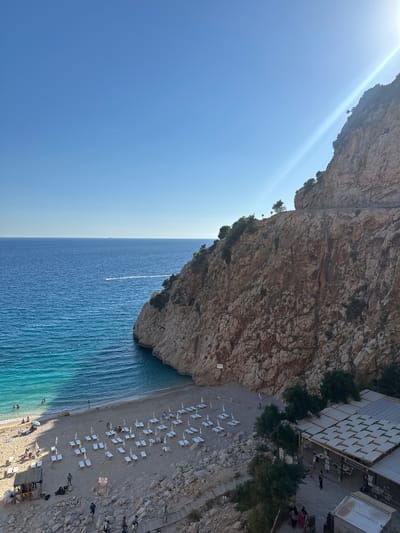A picturesque beach cove in Kaş, Turkey, is captured from an elevated perspective, likely during the late afternoon or early evening, given the soft light and long shadows cast on the sand. The scene is dominated by a long, narrow stretch of sandy-pebble beach situated at the base of immense, rugged cliffs on the right side of the frame.

Numerous white sun loungers are neatly arranged across the beach, with several people relaxing or sunbathing. A few individuals are visible in the clear, turquoise-to-deep-blue waters of the sea, while others walk along the shoreline. Further up the beach, to the right, are several multi-tiered beach structures with white fabric awnings, suggesting beach bars or restaurants. Visible text on one structure reads "LAS ROCAS BEACH..." (partially obscured), and another clearly displays "LA GELATERIA," indicating an ice cream parlor. Wooden stairs lead up from the beach to these establishments.

The vast expanse of the Mediterranean Sea stretches out to the left, its surface rippling with gentle waves. A small white boat is visible in the distance, leaving a faint white wake behind it. The sky above is a clear, bright blue, with a strong sun glare originating from the upper left. The towering cliffs are composed of light brown and grey rock, with sparse green vegetation clinging to their steep sides. A small red and white warning sign is also present on the beach. The overall atmosphere conveys serene coastal relaxation and natural beauty.