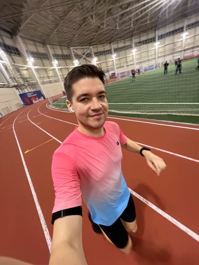A man smiles in a selfie while jogging on an indoor track. He wears a pink and blue ombre long-sleeved shirt, black leggings, and a fitness tracker. The indoor track is red with white lines, and a green netted area borders the track. Overhead, the structure of the indoor facility is visible with bright lights. Other figures are dimly visible on the opposite side of the track.
