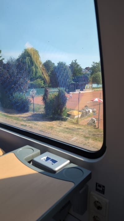 The image shows a view from inside a train, looking out of the window. The window is large and reflects the interior of the train. Outside, a tennis court with red clay is visible. There are several people on the court, seemingly playing tennis. Some trees and a building are also visible in the background. The weather appears to be sunny and clear, suggesting it is daytime.
The interior of the train shows a table, an electrical outlet, and a blue sign with a white symbol. The sign appears to have text that is partially obscured. The view outside the window is partially obscured by reflections on the glass. The overall scene depicts a sunny day with outdoor recreational activity. The location is implied to be Offenburg, Germany.
