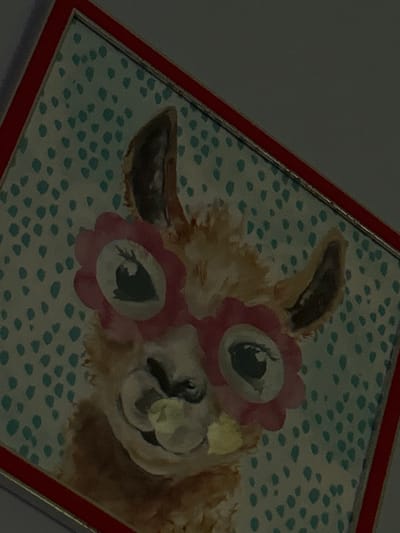 The image is a close-up of a framed painting of a llama wearing pink, flower-shaped glasses. The llama is smiling and appears to be chewing on something. The background of the painting is a teal color with a pattern of small, white dots. The frame is red and gold. The image is likely from a home or hotel room in Montañita, Ecuador, based on the coordinates provided. The coordinates correspond to a location on the coast near the center of Montañita.