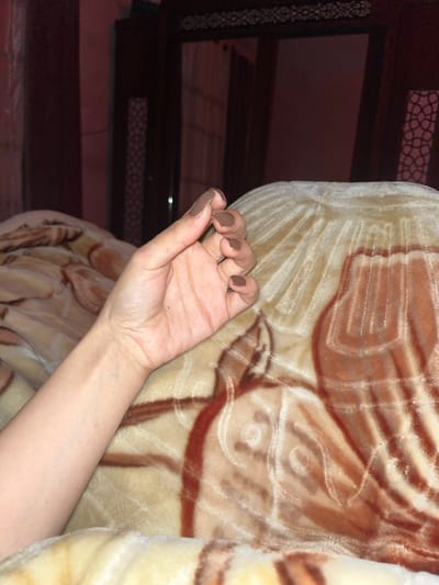 A person's left hand is extended with fingers slightly curled, displaying dark brown nail polish. The arm extends from the hand, resting against a beige and brown patterned blanket. To the left, a dark brown wooden dresser with decorative latticework is partially visible. The overall lighting is dim.
