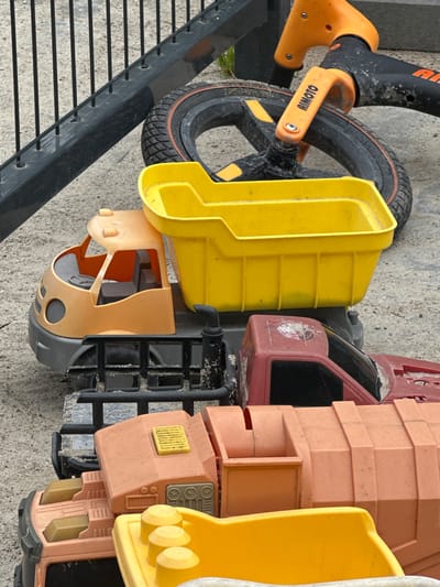 The image shows several toy trucks and a child's scooter on a paved surface. The toys are dusty and appear to have been left outdoors. One toy truck is orange and grey, with a yellow dump bed. Another is red. A child's scooter with orange handlebars and a black frame is also visible. The background includes a black metal fence and some greenery. The overall scene suggests an outdoor play area or a yard. The location is identified as Malinovka, Russia.