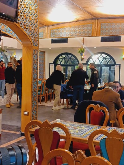 The image captures an indoor scene within an establishment located in Dummar Municipality, Syria. The environment features ornate Middle Eastern architectural design, including an arched wooden doorway with intricate geometric lattice-work in gold and light blue, extending onto adjacent walls. The ceiling is paneled with warm, diffused lighting. White walls are visible in the background, punctuated by multiple large arched windows with dark frames and internal grid patterns, suggesting an exterior view or adjoining room.

The flooring consists of patterned carpets and potentially tiled sections. Furniture includes wooden tables with colorful tiled mosaic tops and wooden chairs featuring carved details and upholstered seats in red and blue. Additional dark blue armchairs are visible further back. A black Sony camera lens rests on the floor in the lower-left foreground.

Approximately twelve individuals, mostly men, are present. In the midground, three men are standing and conversing: one with a light head covering and dark clothing, another with grey hair in a dark jacket, and a third in a dark leather-style jacket with a white stripe on the sleeve. To the left, near the arched doorway, several men are standing, including one in a red shirt with a beard, and another in a dark coat. In the immediate foreground on the right, a man in a brown jacket is seated at a tiled table, where a small white cup is placed. The overall scene suggests a casual social gathering or dining environment.