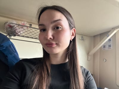 The image depicts a female individual within a train compartment, situated in Usolye-Sibirskoye, Russia. The subject, with long, straight, dark brown hair and a fair complexion, occupies the foreground, looking directly at the camera. She is wearing a black long-sleeved garment. Her right ear features a small hoop earring with a clear stone on the lobe and a gold-colored hoop earring on the helix. The background reveals beige-paneled walls typical of a passenger train. Above the subject's left shoulder, a metal mesh rack holds two clear plastic bags containing indistinct items. To her right, a set of metal coat hooks is affixed to the wall. Further right, an articulated wall-mounted mechanism, possibly for a fold-down table, is visible, along with two unreadable QR code decals. A blue backpack is partially visible on the far left side of the frame.
