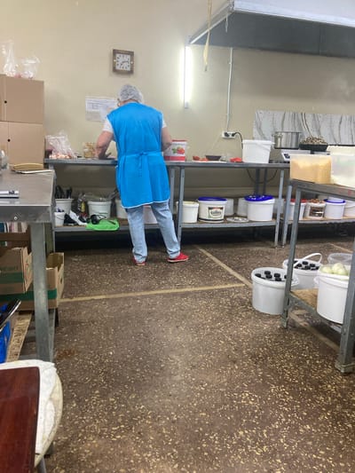 The image shows a person wearing a blue apron and hairnet working in a commercial kitchen, likely in Balakovo, Russia. The kitchen appears functional with stainless steel work surfaces and shelving units. Various food containers, buckets, and ingredients are visible on the shelves and floor. A clock on the wall suggests it is late afternoon. The floor is made of speckled concrete. There is no indication of the specific location in Balakovo, only that it is a kitchen environment.