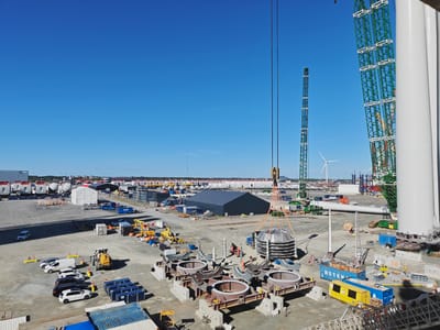 This is an outdoor, daytime scene at a port or industrial facility, likely in Esbjerg, Denmark, given the context. The clear blue sky suggests fair weather. The scene is dominated by the construction of large industrial components, possibly for wind turbines, as indicated by the presence of what appear to be tower sections, blades, and cranes.
The main subjects are the various components and machinery. In the foreground, several large, circular metal structures are laid out on concrete bases, each with intricate gear-like mechanisms. A heavy-duty crane, suspended by thick ropes, is in the process of lifting a large, multi-layered circular object. Several vehicles, including cars, vans, and a yellow forklift, are parked or positioned around the site. In the background, a massive green lattice crane structure is partially visible, as are tall, white tower sections. Numerous other structures, tents, and containers are scattered across the expansive, dusty ground.
There are a few individuals visible, appearing to be workers in orange vests or safety gear, managing the operations. One person is visible near the forklift, and others are near the components being prepared.
Notable details include the sheer scale of the construction elements and the advanced machinery. The bright sunlight creates distinct shadows, indicating it's likely midday. The overall impression is one of a busy, industrial work site engaged in a significant construction project.
Visible text includes "STEN" on a blue shipping container and "ESTE" on a yellow container.