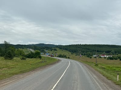 The image depicts a winding asphalt road in Krilovo, Russia, under a cloudy sky. The road is flanked by grassy verges and a line of trees. In the distance, rolling hills covered with a dense forest create a lush green backdrop. Several vehicles are visible on the road, including a red and white truck. Some buildings, appearing to be houses or small structures, are scattered across the landscape in the distance, some with red roofs. Road signs and utility poles are also present along the roadside. The overall impression is of a rural, somewhat overcast day in a Russian countryside setting.