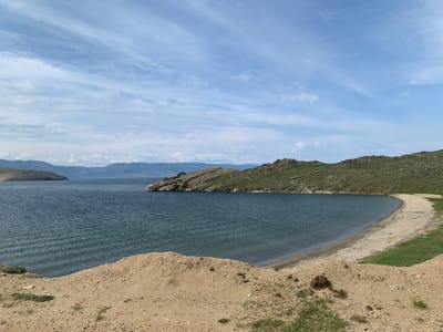 A wide shot captures a serene natural landscape in Sakhiurta, Russia. In the foreground, a sandy bank slopes gently downwards towards a body of dark blue water. The water is calm, with subtle ripples disturbing its surface, and it stretches to meet a distant shoreline. Beyond the immediate water, rugged, green-covered hills rise up, interspersed with rocky outcrops. These hills form a prominent natural barrier, with some parts appearing barren and rocky, while others are densely vegetated. In the far distance, a hazy, blue mountain range forms the horizon. The sky above is a vast expanse of pale blue, adorned with wispy, white clouds that stretch across the frame. The overall scene conveys a sense of tranquility and untouched natural beauty.