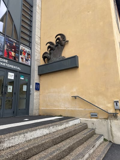 The image depicts the exterior of a public or institutional building in Le Puy-en-Velay, France, specifically associated with a "Médiathèque" and "Départemental" services, indicated by banners on the facade, and an "IURP" logo on the entrance doors.
The scene is dominated by two distinct architectural styles: a modern, dark grey panelled facade with large glass windows on the left, and an older, light yellow stucco wall on the right, exhibiting some weathering and discoloration. The main entrance, located on the left facade, consists of double grey-framed glass doors. Above these doors are banners advertising "THEATRE", "MÉDIATHÈQUE", and "HISTOIRE DÉPARTEMENTAL".
A prominent dark-colored, possibly bronze, abstract sculpture of three stylized roosters (coqs) is mounted on the yellow stucco wall, positioned above a dark, empty rectangular plaque. The roosters feature flowing, curvilinear forms suggesting their combs and tail feathers.
Access to the building is via a set of exposed aggregate concrete stairs with dark treads and lighter risers, leading upwards from the foreground. The top surface of these steps is marked with black and white stripes. A black metal handrail is affixed to the yellow wall alongside the right side of the stairs. Below the handrail, a small grey rectangular vent or grate is visible at ground level. Further right, a grey wall-mounted utility box or service panel is present. A blue informational or historical plaque is affixed to the wall near the double doors. The immediate ground in the foreground consists of dark pavement with a white striped section.
No individuals are present in the scene, and no explicit actions or interactions are depicted. The lighting is even, suggesting an overcast day.