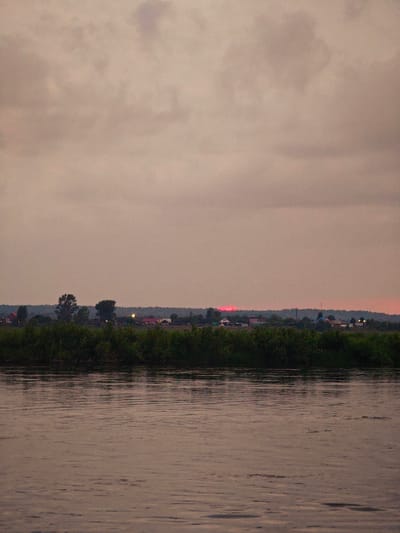 The image shows a wide, calm river in the foreground, with ripples on its surface reflecting the dusky sky. A dense line of green foliage forms the bank on the opposite side of the river. Behind the foliage, a low-lying village or settlement is visible with scattered houses and some trees. The sky above is overcast with muted, pastel tones, suggesting either dawn or dusk. In the distance, a faint, horizontal band of red light is visible near the horizon, likely indicating the setting or rising sun partially obscured by clouds or landscape. The overall atmosphere is serene and somewhat melancholic. The village appears to be in a rural area, consistent with the location of Kazachinskoye, Russia, which is situated along the Angara River and surrounded by natural landscapes. There are no people or specific objects that would allow for further identification of the scene's activities or events.