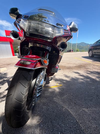 This is a low-angle, eye-level shot of the rear of a dark red Honda motorcycle parked on a paved surface in Sotonići, Montenegro. The motorcycle's large rear tire is prominently featured in the foreground, casting a shadow on the asphalt. The motorcycle's chrome accents, taillight, and exhaust pipes are visible. In the background, a black car is parked to the right, and a building with red and white elements is visible to the left. Beyond the parking area, mountains and a clear blue sky with some wispy clouds are depicted. The scene appears to be a sunny day, and the lighting suggests it's midday. There are no people or animals visible in the frame.