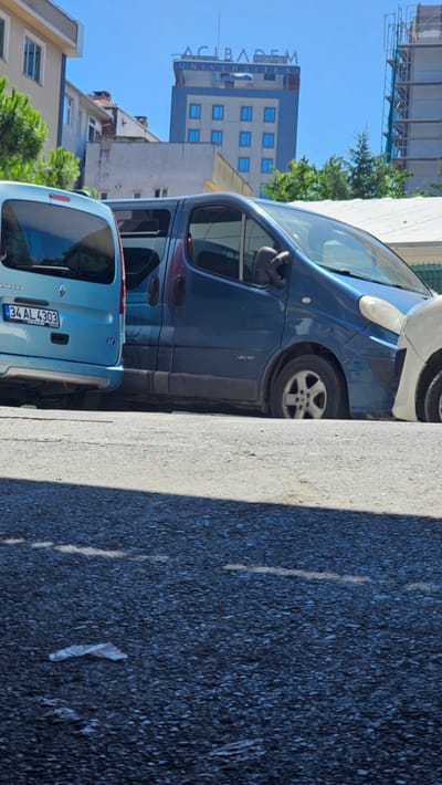 The image shows a street scene in Ataşehir, Turkey, with several vehicles parked. In the foreground, the asphalt road is visible, with some debris on the ground. In the middle ground, two blue vans are parked. The van on the left is a light blue Renault Kangoo, with a license plate reading "34 AL 4303". The van on the right is a darker blue, likely a Renault Trafic, with the model designation "dCi 115" visible on its side. In the background, a tall, modern building with the "ACIBADEM UNIVERSITESİ" signage is prominent. Construction scaffolding is also visible on another building to the right. The sky is clear and blue, indicating a sunny day. The scene appears to be in an urban environment.