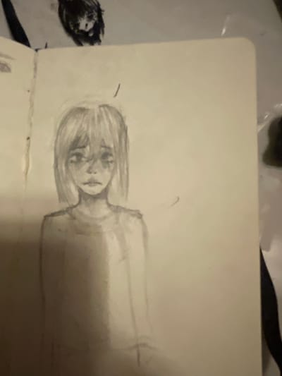 A pencil sketch on a piece of paper depicts a person lying down. The subject is drawn from the shoulders up, with their head turned to the side and their eyes closed. They have short, wispy hair. The sketch is rendered in a soft, shaded style, giving it a somewhat ethereal quality. The background is blank, suggesting the drawing is in a sketchbook or on a loose sheet of paper. To the left of the sketch, a dark, fuzzy object appears to be a crumpled piece of fabric or paper. The overall impression is one of quiet repose or perhaps sleep. The sketch is a simple and intimate portrayal of a person.