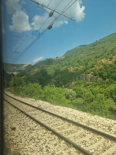The image shows a view from a moving train through a window. In the foreground, train tracks are visible, laid on a bed of light-colored gravel. The tracks curve to the left. Overhead, electrical wires for the train are suspended from poles, with insulators visible. In the middle ground and background, a lush green hillside rises steeply, covered in dense trees and vegetation. A narrow, dark river or stream can be seen winding through the trees at the base of the hill. The sky is a clear, bright blue with a few white clouds. The scene is illuminated by bright daylight, casting shadows on the hillside. The photo appears to be taken in a natural, mountainous environment. Based on the context provided, this scene is located in the city of Milanovo, Bulgaria.