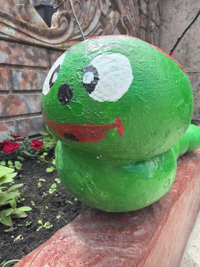 The image shows a close-up of a large, bright green, round object with painted features, resembling a cartoon character or a decorative garden ornament. The object has a white patch with a black pupil for an eye, and a red stripe with a black dot underneath it, suggesting a face. It is positioned on a textured surface that appears to be a tiled or paved area.

Behind the green object, another similar green, rounded form is visible, and further back, a reddish-brown, textured surface curves around. The ground next to the green object is dark soil with small green plants and a few red flowers.

A thin black wire extends from the top left of the green object, disappearing out of frame. The lighting suggests it is daytime, and the overall scene appears to be outdoors, possibly in a garden or yard. The art style of the green object is playful and somewhat simplistic. There is no visible text in the image. The location context provided is Velyki Osniaky, Ukraine.