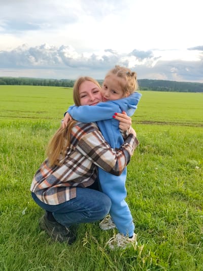 A young girl hugs a woman in a field. The woman is crouching. The woman is wearing jeans and a plaid shirt. The girl is wearing a blue hoodie and pants. The background is a green field and a cloudy sky. The location is Горняк, Russia.