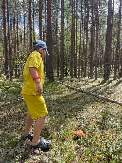 In Dom Otdykha, Russia, a young person wearing a bright yellow t-shirt and shorts, a blue baseball cap, and dark clogs with charms, is walking through a dense pine forest. The person is observed from behind and to the side, with their left arm adorned with a bright pink bracelet. They are looking down at a single, brown mushroom on the forest floor. The ground is covered in moss, small green plants, and scattered pine needles. Sunlight filters through the tall pine trees, creating dappled shadows on the forest floor. A fallen log lies horizontally across the scene in the middle ground. The overall environment is one of a natural, wooded area.