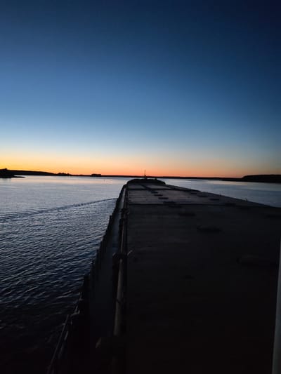 The image depicts a tranquil, wide body of water, likely a bay or gulf, at dusk. The sky transitions from a deep, clear blue at the top to a soft gradient of orange and yellow near the horizon, indicating the sun has recently set or is about to rise. A dark, landmass with silhouetted trees lines the distant horizon.
In the foreground, a long, dark, flat-topped structure extends into the water, creating a pier or a section of a larger vessel. The surface of this structure is textured and appears to be made of concrete or a similar material, with a pattern of rectangular indentations. The water to the left of this structure is rippled, with a faint wake suggesting movement. The water is dark, reflecting the dimming sky. A few small, distant lights are visible along the shoreline. The overall mood is serene and peaceful, characteristic of a quiet evening by the water. The location is the city of Irdomatka, Russia.