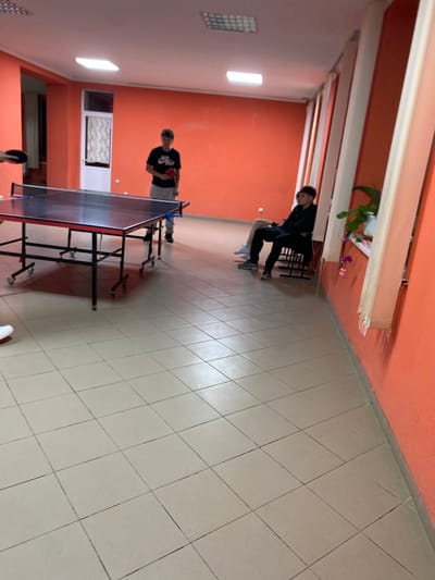 This media file captures an indoor scene at what appears to be a community center or recreational facility in Negrești, Moldova. The subjects are two young individuals, likely teenage boys, engaged in a game of table tennis. One boy is standing on the left side of the table, wearing a black t-shirt with white text and grey shorts. He is holding a table tennis paddle and appears to be in the process of playing. The other boy is positioned on the opposite side of the table, also holding a paddle.
The setting is a long room with orange-painted walls and a tiled floor. The room is lit by artificial lights, suggesting it is either evening or an indoor space without natural light access. A ping pong table dominates the center of the frame. In the background, there are windows with blinds, and some plants on a shelf near the wall. The overall atmosphere seems casual and recreational, with the focus on the game. There are no strong indicators of time of day or weather conditions, as it is an indoor shot. The emotion conveyed is one of concentration and mild activity related to the game. The text visible on the t-shirt of the boy on the left appears to be the Nike logo with "NIKE" written beneath it.