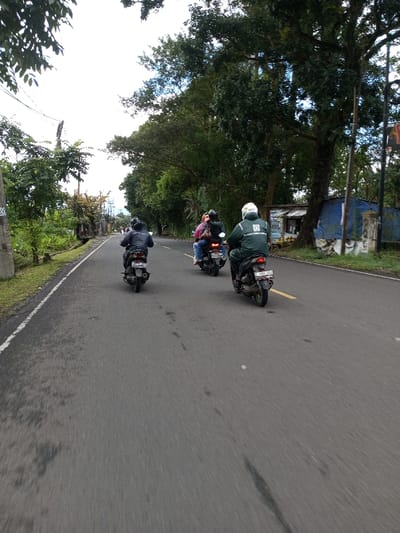 The image depicts a view along an asphalt road in Serpang Shalli, Bhutan, under an overcast sky. Three individuals are riding motorcycles away from the viewer. From left to right: a solo rider in a dark jacket and dark helmet on a dark scooter; a second scooter with two occupants, the rear passenger wearing a pink top and dark helmet; and a solo rider in a green jacket with a white logo on the back, wearing a white helmet, on a dark scooter with license plate "F 5885 VLC". The road surface shows dark stains and is bordered by a white line on the left. The left side of the road features a grassy verge, a concrete utility pole with "380/200" and "02" visible, and dense green foliage. The right side of the road is lined with large trees, under which a makeshift structure of corrugated metal and blue tarpaulin is situated, possibly a stall, with light-colored objects visible in front. A concrete pillar stands next to this structure.