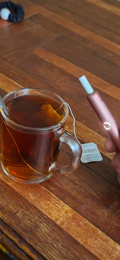 A close-up shot captures a warm beverage in a clear glass mug, accompanied by a smoking device. The mug, filled with dark tea and a tea bag, sits on a wooden surface. A rose-gold colored smoking device, with a white tip, is held by a hand in the right side of the frame, positioned above the mug. A small white tag with the word "Carlton" and "PREMIUM QUALITY" is attached to the mug. In the upper left corner, a black hair tie with a white bead is visible. The background consists of a wooden table with a distinct grain pattern. The image does not contain any elements that would allow for the identification of its specific location in Shakhtarsk, Ukraine, or any other city.