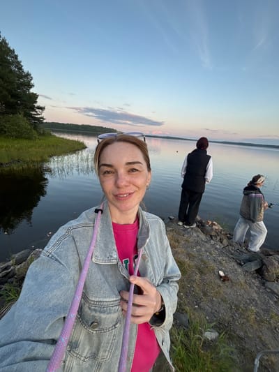 A woman is taking a selfie on the shore of a lake at dusk, with two people fishing in the background. The woman in the foreground is wearing a denim jacket over a pink shirt and has sunglasses on her head. The lake is calm and reflects the soft colors of the sky. The shore is rocky and covered with grass and some vegetation. The two people in the background are standing at the edge of the water, casting their fishing lines. The overall scene suggests a peaceful evening spent outdoors.