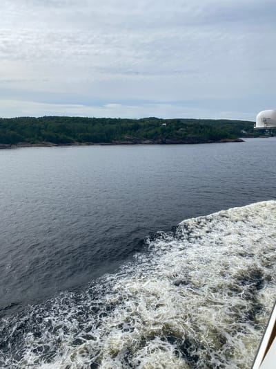 The image captures a view from a boat sailing in the waters off the coast of Larvik, Norway. The dark, choppy water, disturbed by the boat's wake, dominates the foreground. The wake is a frothy white, swirling and churning as it recedes. In the middle ground, a rocky shoreline lined with dense, green coniferous trees stretches across the frame. A few scattered houses are visible amongst the trees, suggesting a sparsely populated coastal area. The background is filled with a pale, cloudy sky, with wisps of white clouds creating a textured pattern. A white dome-shaped object, possibly part of the boat's superstructure, is visible in the upper right corner. The overall scene depicts a natural, serene coastal landscape under an overcast sky.