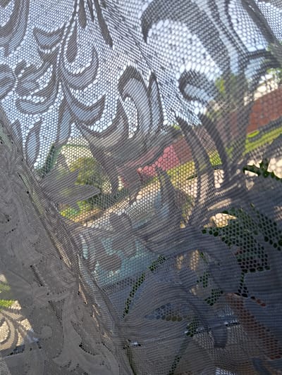 The image displays an interior perspective looking through a light grey, patterned lace curtain towards an outdoor scene in the city of Teykovo, Russia. The lace curtain, sharply in focus, exhibits a repeating ornate scrollwork and floral motif composed of denser fabric against a fine hexagonal mesh. The curtain's surface shows slight undulations and creases. Beyond the semi-transparent lace, the exterior environment is visible in soft focus under what appears to be a bright, possibly overcast, daytime sky. Elements discernible in the background include green foliage consistent with trees or bushes, a section of a reddish-brown tiled roof from a building, and hints of a light-colored path or road. No individuals, specific actions, or interactions are depicted within the frame.