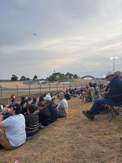 A crowd watches an event outdoors. People sit in folding chairs and on the ground, facing toward a racetrack. A helicopter is visible in the sky. A Dunlop arch is visible on the track in the distance. Trees line the track. The sky is cloudy.