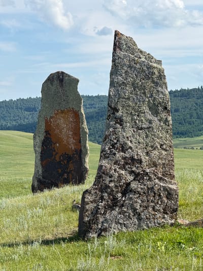 Two ancient, weathered standing stones are positioned in a grassy field under a partly cloudy sky. The stone in the foreground is larger and rougher, with a jagged top edge and covered in lichen. The stone behind it is taller and slimmer, with a smoother surface that displays patches of orange and dark coloration, likely lichen or mineral staining. The field is covered in green grass with some lighter-colored grasses and small wildflowers. In the background, a densely forested hillside slopes gently upward, with a lighter green hill visible further in the distance. The sky is blue with scattered white clouds. The location is described as being in the city of Сафронов, Russia, which is likely referring to an archaeological site or natural landmark within or near that area. There are no people or animals visible in the image, nor any discernible actions or interactions. The scene is static and appears to be a historical or natural landscape.