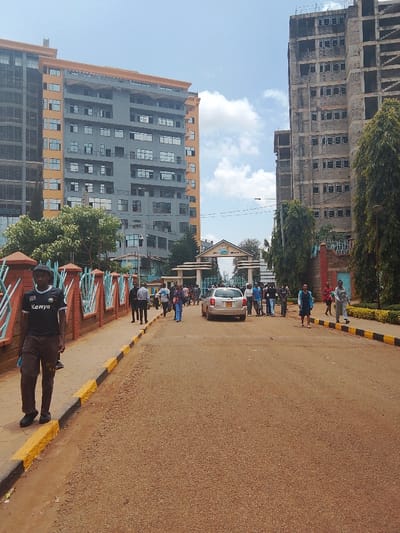This image depicts a daytime street scene in Karingani ward, Kenya, characterized by mixed urban development. In the foreground, an unpaved or lightly paved brown road occupies the center, flanked by sidewalks with yellow and black painted curbs.
On the left side, a male individual, appearing adult, walks on the sidewalk. He is wearing a black short-sleeved shirt with "Kenya" text in white on the front, dark knee-length shorts or trousers, and dark shoes. He holds a light blue item in his left hand. A brick wall with blue metal decorative elements borders the sidewalk to his left. Behind him and further down the street, a multi-story building, partially constructed or recently completed, stands prominently. It features a grey facade with numerous windows and an orange-brown top and side trim.
In the middle distance, a light-colored sedan or hatchback vehicle is positioned on the road, facing away from the viewer, with its rear visible. Numerous pedestrians, approximately 15 to 20 individuals, are present on the road and sidewalks, predominantly congregating near an arched white gateway that bears text, partially legible as "HILL INSTITUTE." Beyond this gateway, another building is visible.
On the right side of the road, a tall, unfinished concrete building structure, appearing to be a skeletal framework, stands against the sky. Evergreen trees line the sidewalk on this side. The sky above is clear with scattered white clouds.