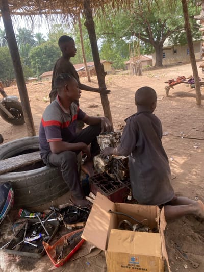 This image depicts an outdoor motorcycle repair scene in Okura, Nigeria, under a rudimentary thatched shelter supported by wooden poles. Three males are present, engaged with mechanical components on a dry, reddish-brown dirt ground.
In the foreground, an adult male, likely a mechanic, sits on two stacked dark tires topped with a wooden plank. He wears a grey and red-striped polo shirt and dark trousers, with grease visible on his hands. Directly in front of him are disassembled motorcycle engine parts, including a cylinder head and crankcase, resting on a red plastic crate and the ground. To his right, a young male child, estimated 7-10 years old, kneels with his back partially to the camera, wearing a dark, dirt-stained shirt and interacting with the engine components. Behind the seated adult, a taller young male, approximately 12-16 years old, stands, wearing a dark top, with his right hand resting on one of the support poles.
Various tools, including wrenches and smaller metallic parts, are scattered in a red plastic tray and directly on the ground. An open brown cardboard box, labeled "JEZCO OIL ROSY SPECIAL," sits in the immediate foreground, containing additional mechanical parts. A portion of a motorcycle frame and front wheel is visible on the far left.
The background reveals sparse vegetation, mature trees, and several residential structures, including simple mud-walled buildings with thatched roofs and a lighter-colored building with windows, indicating a rural or semi-urban environment under clear daylight conditions.