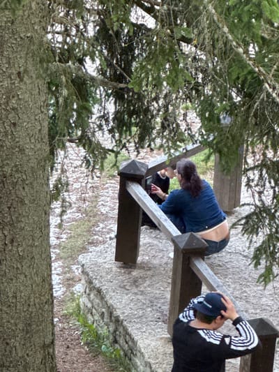 In Keila-Joa alevik, Estonia, a man wearing a dark baseball cap and a black and white striped long-sleeved shirt is standing on a stone pathway, adjusting his cap. Nearby, a woman in a blue denim jacket and jeans is seated on the pathway. She is looking at a black object in her hands, possibly a phone. Another person is partially visible behind her, also appearing to be looking at the object. The scene is outdoors, with a large tree on the left and coniferous branches overhead. The pathway is bordered by a wooden railing. The ground is covered with gravel and some scattered white debris, possibly snow or ice.