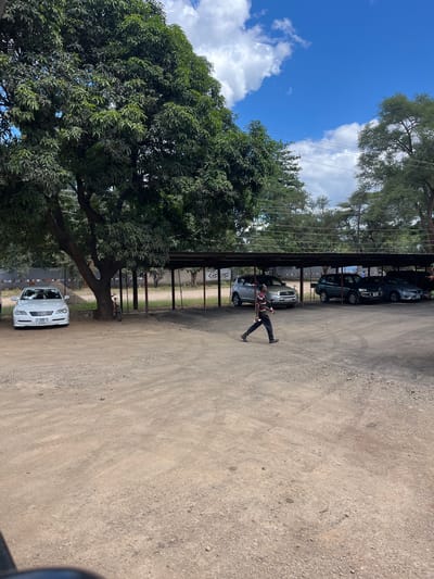 The image depicts an outdoor scene in Livingstone, Zambia. A person is walking under a metal awning, where several cars are parked. There is a large tree to the left side of the image, with a car parked next to it. A bicycle is resting against the tree. The ground is gravel. The sky is visible with some clouds.