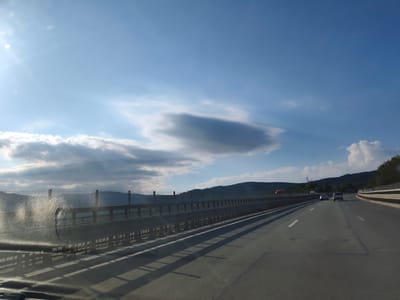 The image captures a view from a vehicle traveling on a highway in Vrachesh, Bulgaria. The highway has multiple lanes and is bordered by a metal guardrail and a fence. The sky is a bright blue with scattered clouds, and the sun is visible in the upper left corner, casting lens flares and creating a bright sheen on the vehicle's windshield. In the distance, rolling hills covered in vegetation can be seen. Several cars are on the highway, and shadows from the guardrail stretch across the asphalt. The overall scene suggests a scenic drive on a sunny day.