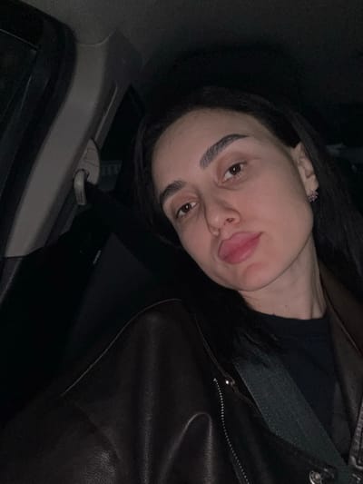 A person with brown hair and eyebrows, wearing a dark brown leather jacket and a black shirt, is seen from a low angle inside a car. They are wearing a seatbelt and sticking out their tongue. The car door and handle are visible. The image is dimly lit, making the colors appear muted.