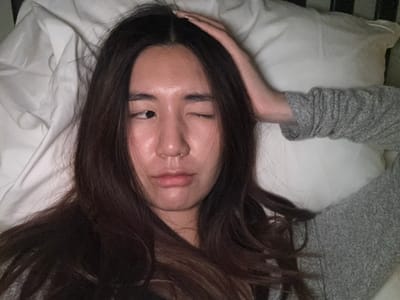 A female individual is depicted from a high angle, lying supine on white bedding in an indoor setting. Her long, dark brown hair, parted down the middle, is spread across a white pillow. She presents a winking expression; her right eye is closed, and her left eye is partially open, looking towards the right side of the frame. Her lips are slightly parted, and a small silver ring is visible in her right nostril. An arm, covered by a grey, textured long-sleeved garment, extends from the right side of the frame with its hand resting gently on the subject's head. Crinkled white sheets or duvet covers are visible in the left foreground, while a dark vertical element, consistent with a bed frame or headboard, is present in the upper right background. The image contains no visual elements indicating a specific geographical location, such as Le Havre, France.